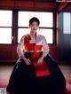 A woman in a red and black hanbok sitting on a red rug.